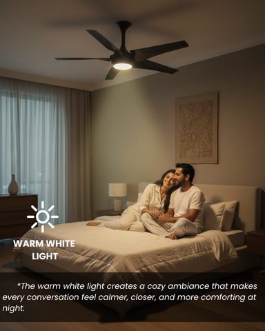 Couple sitting on a bed in a dimly lit room with a ceiling fan and warm white light.